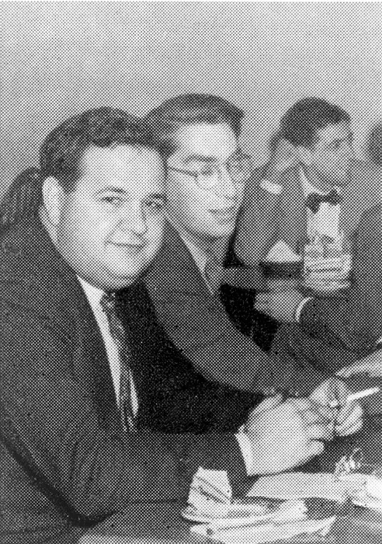 Two individuals seated at a table, smiling and chatting. Others in the background are dressed formally, holding drinks.
