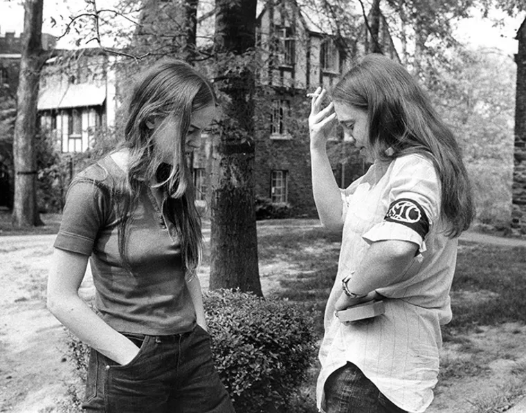Two people stand outside near trees and buildings, one holding a book and looking down, the other touching their head, wearing a black armband.