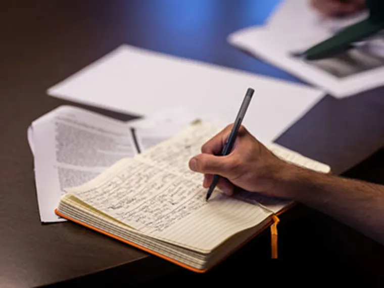 Hand writing in a notebook at a desk, surrounded by paperwork and loose sheets.