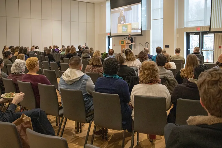 Audience seated in a conference room facing a speaker on stage with a presentation projected on a screen above.