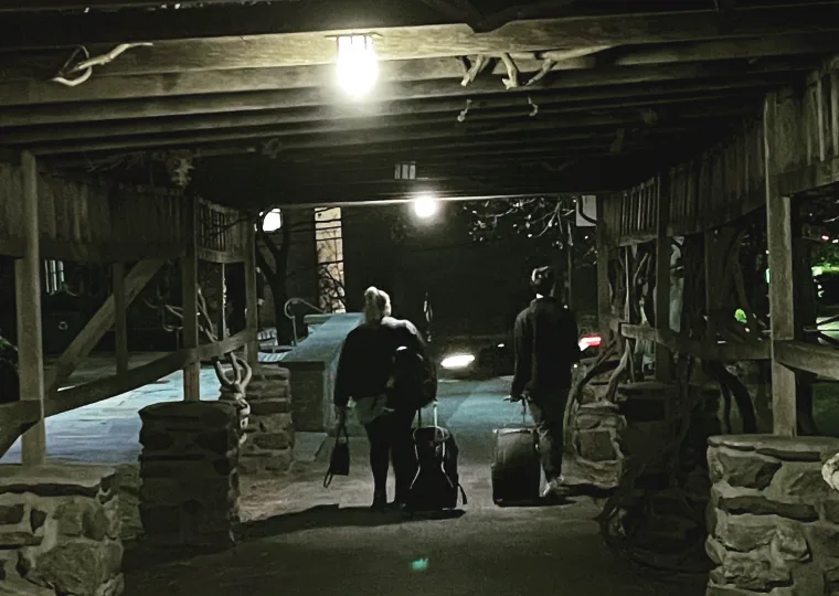 Two people walking under a wooden structure, each pulling wheeled luggage on a dark pathway.