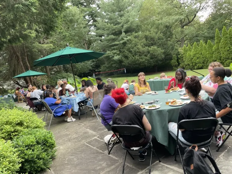 People sitting around tables outdoors, eating and socializing under green umbrellas, with trees and shrubs in the background.