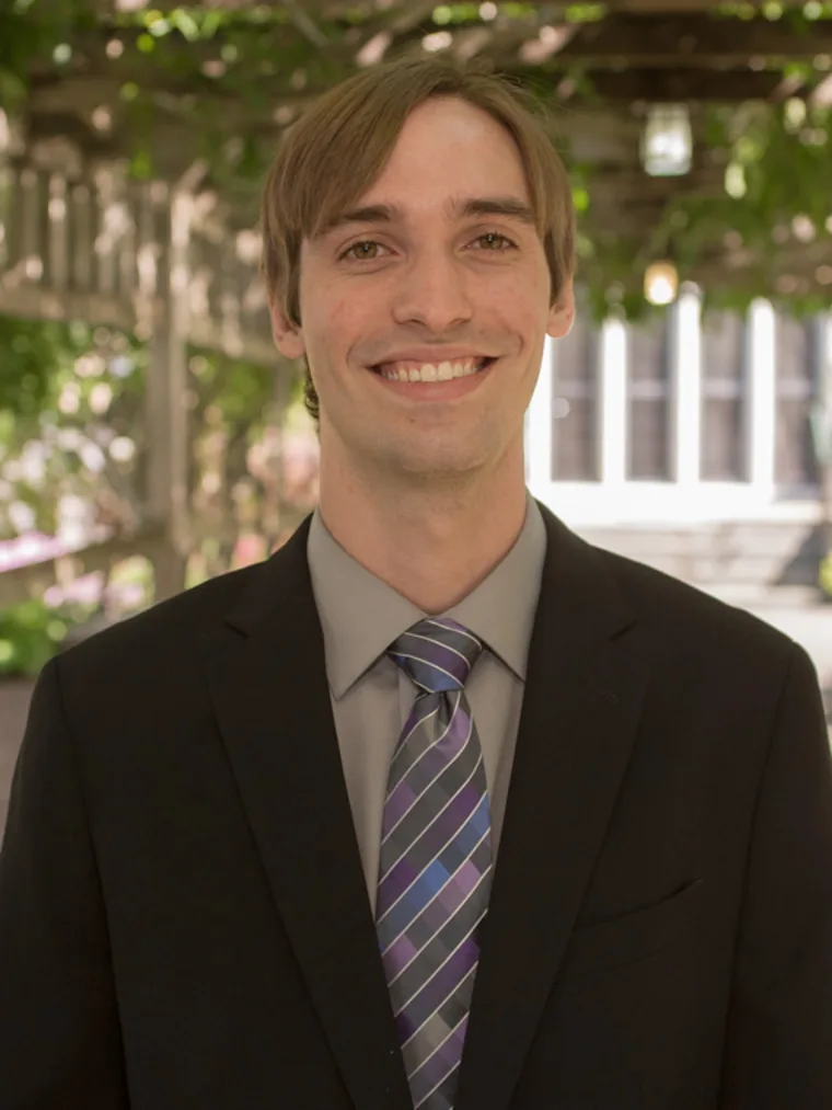 Person in a suit and striped tie smiles outdoors with a wooden pergola and greenery in the background.