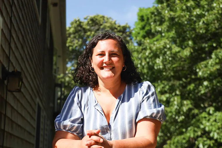 Person with curly hair smiling in a striped shirt, standing outdoors near a house, with trees and blue sky in the background.