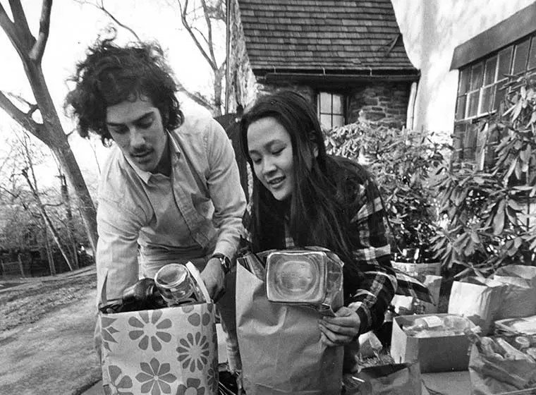Two people sorting groceries into paper bags outdoors, near a house with stone walls and plants in the background.