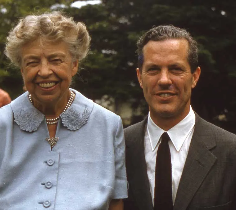 Two individuals are smiling, standing close together outdoors, with trees in the background. One wears a blue outfit with pearls, the other a suit and tie.