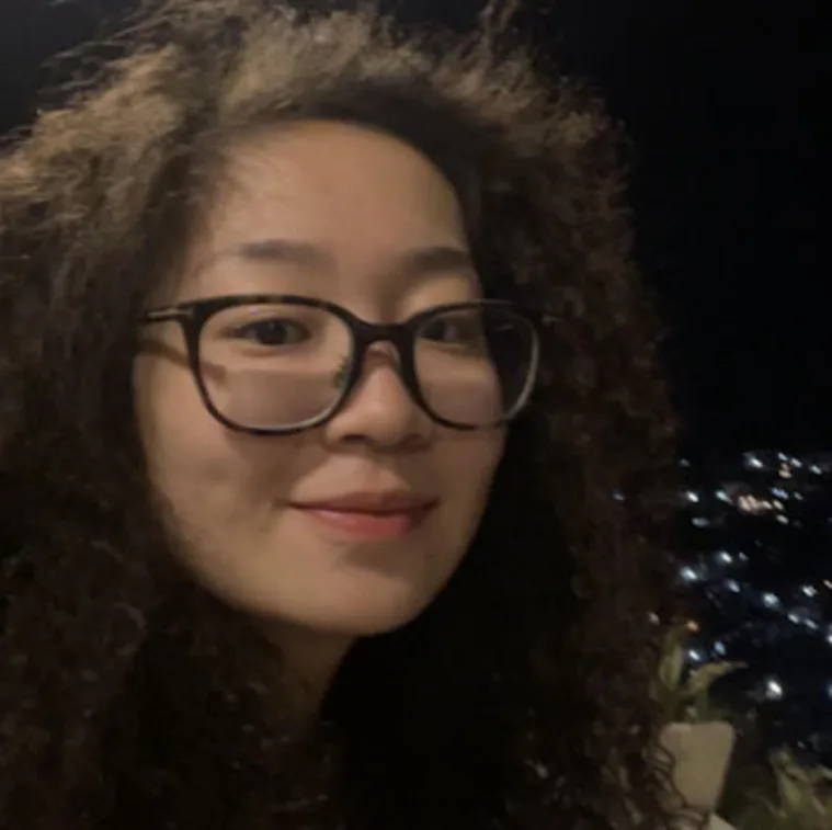 Person with curly hair and glasses smiles at night, with a cityscape in the background.