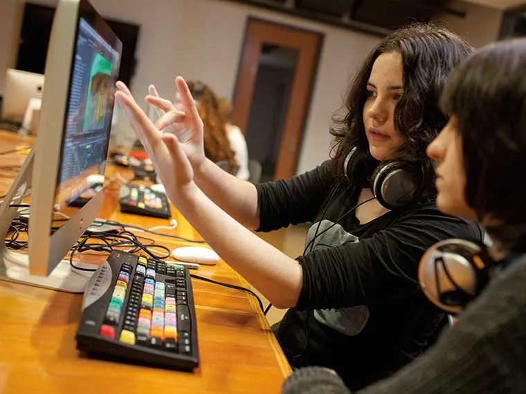 Two people at a desk with computers and headsets, one pointing at the screen while discussing something.