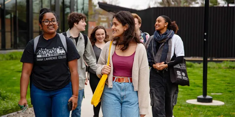 Group of six people walking and smiling outside on a campus pathway, with one wearing a "Sarah Lawrence College" T-shirt.