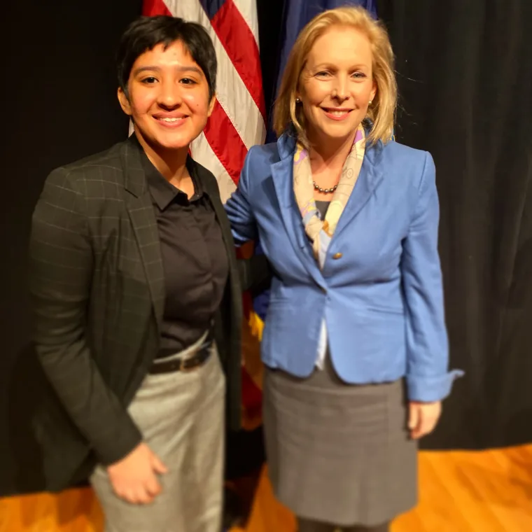 Two people smiling, standing together in front of a U.S. flag. One person wears a dark outfit, the other a blue blazer and scarf.
