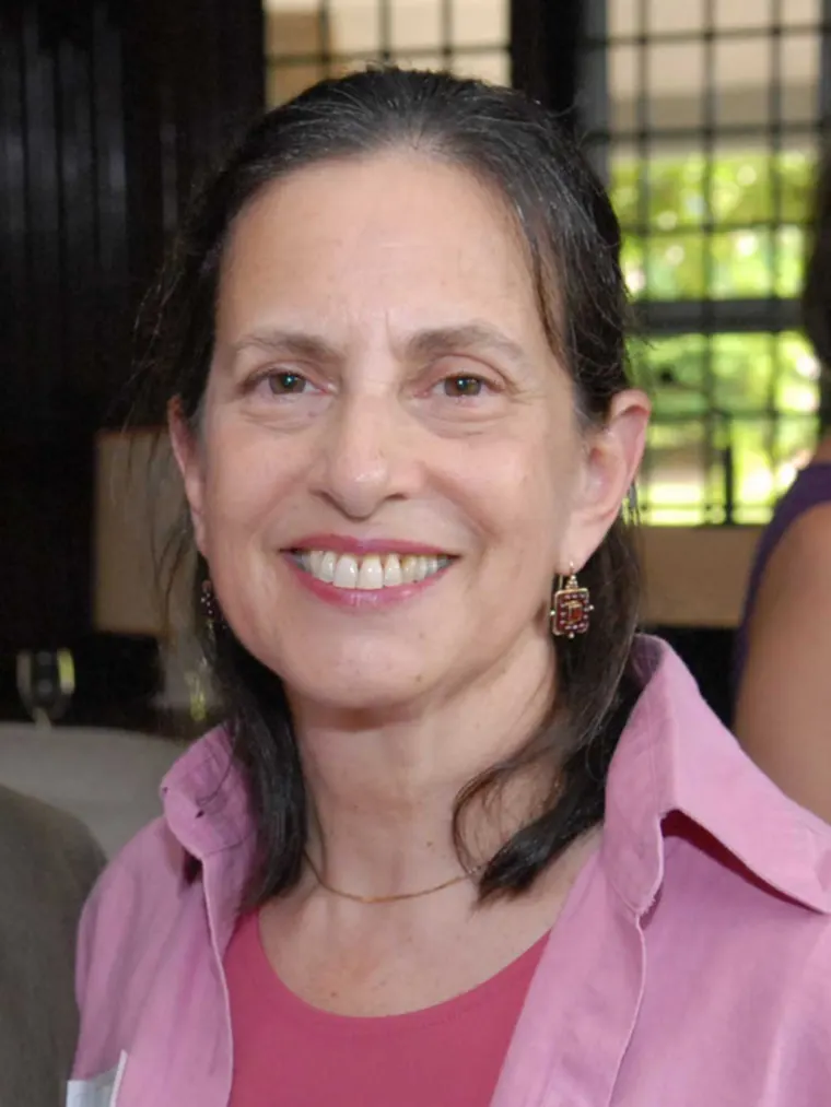 Person smiling, wearing a pink shirt, with long hair and decorative earrings. Blurred indoor background with windows.