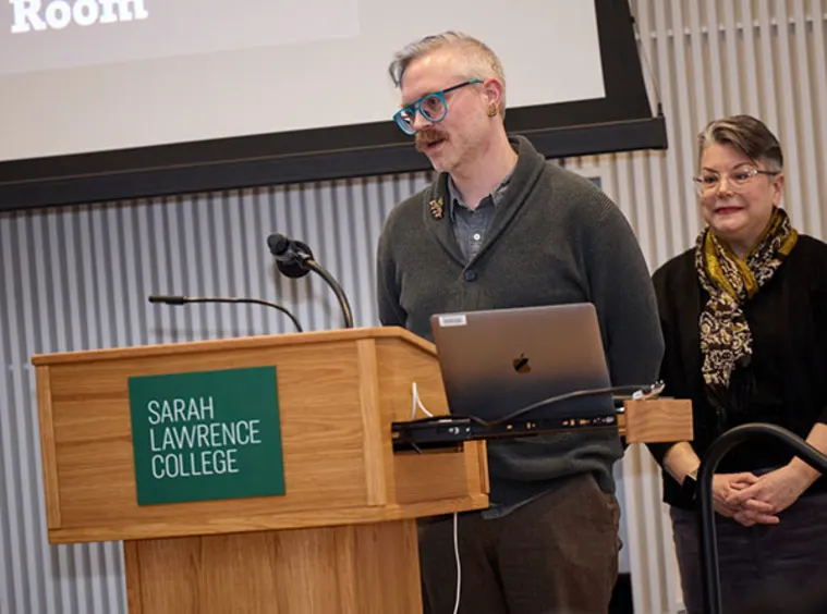 Two individuals speak at a podium with a laptop, under a Sarah Lawrence College sign. A presentation screen is in the background.