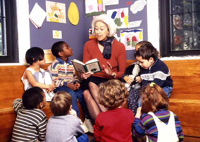 Person reading a book to a group of children sitting on a bench in a classroom with colorful artwork on the walls.