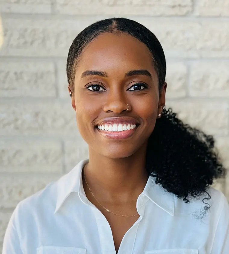 Person smiling in front of a light brick wall, wearing a white collared shirt and a nose ring. Hair is styled in a ponytail.