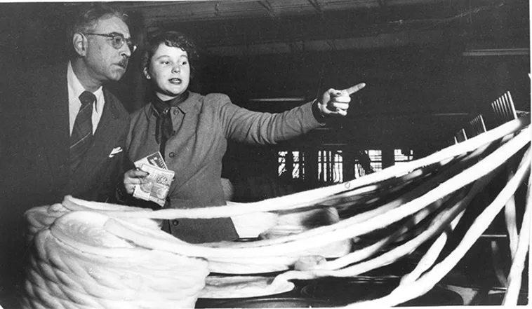 Two people in a textile factory; one pointing while holding yarn, the other observing with interest. Large strands of textile fiber surround them.