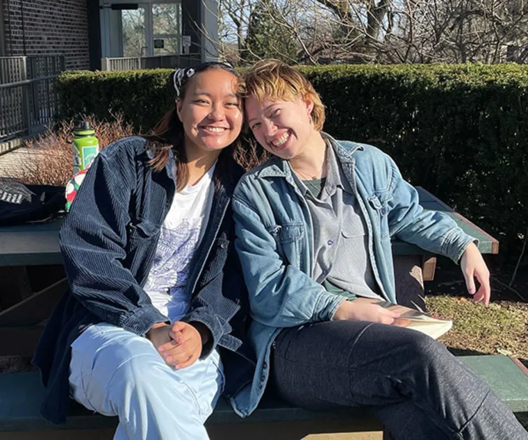 Two people sitting on a bench outdoors, smiling, with one holding a book. They are wearing casual jackets in a sunny setting.