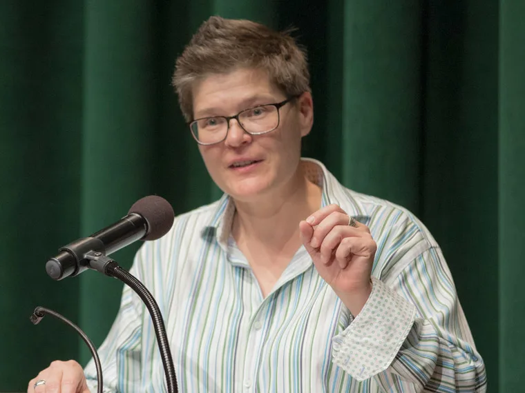Person with short hair and glasses speaking into a microphone. Wearing a striped shirt, gesturing with one hand. Green curtain backdrop.