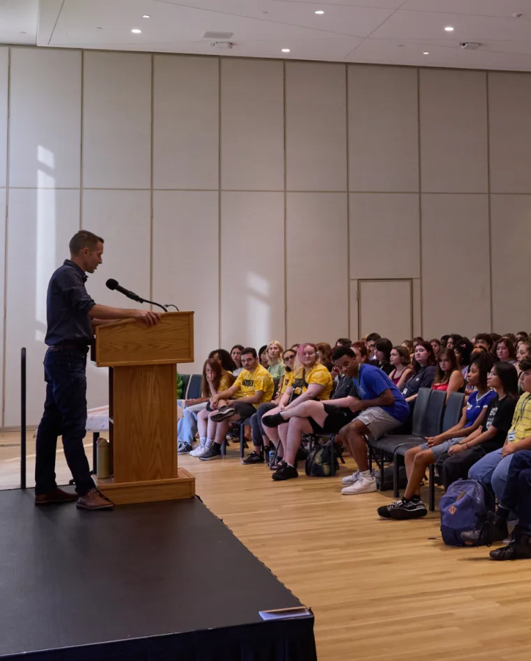 Person speaks at podium to an audience seated in rows, with several attendees wearing matching yellow shirts. Room has beige walls.