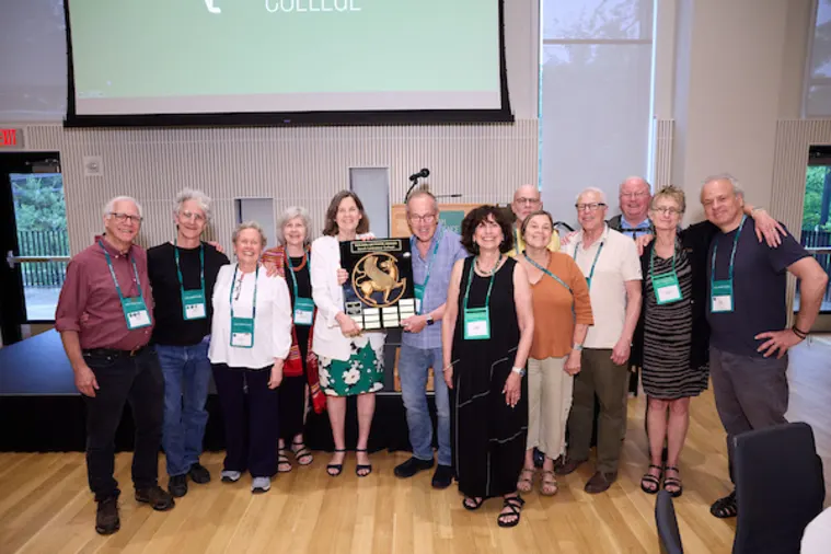 Group of people standing together indoors, some wearing name tags. The person in the center is holding a large plaque.
