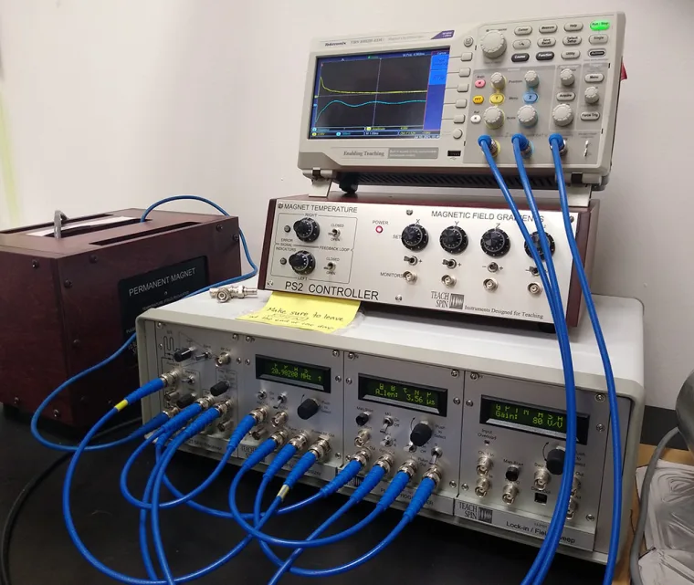 Laboratory equipment setup with connected devices, a monitor displaying waveforms, and a box labeled "Permanent Magnet." Blue cables are attached.