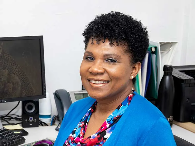Person smiling at a desk with a computer and office supplies in the background. Wearing a blue top and colorful scarf.
