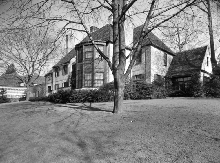 Large brick house with multiple gables and chimneys, surrounded by trees and shrubs, viewed from a yard on a sunny day.