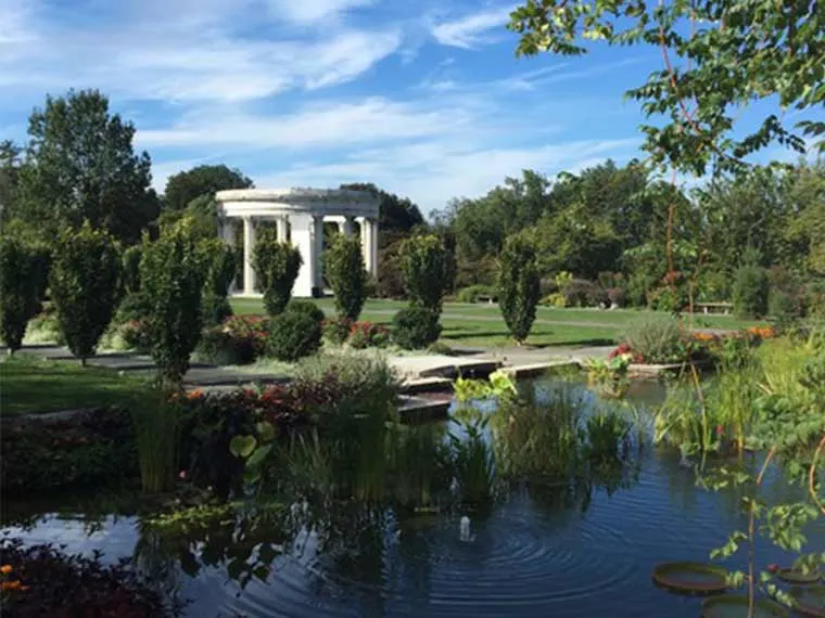 Garden with a pond in the foreground, trees, and a white columned gazebo in the background under a blue sky.