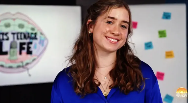 Person smiling, wearing a blue shirt, stands in front of a board with sticky notes and a logo featuring lips and text ending with "LIFE."