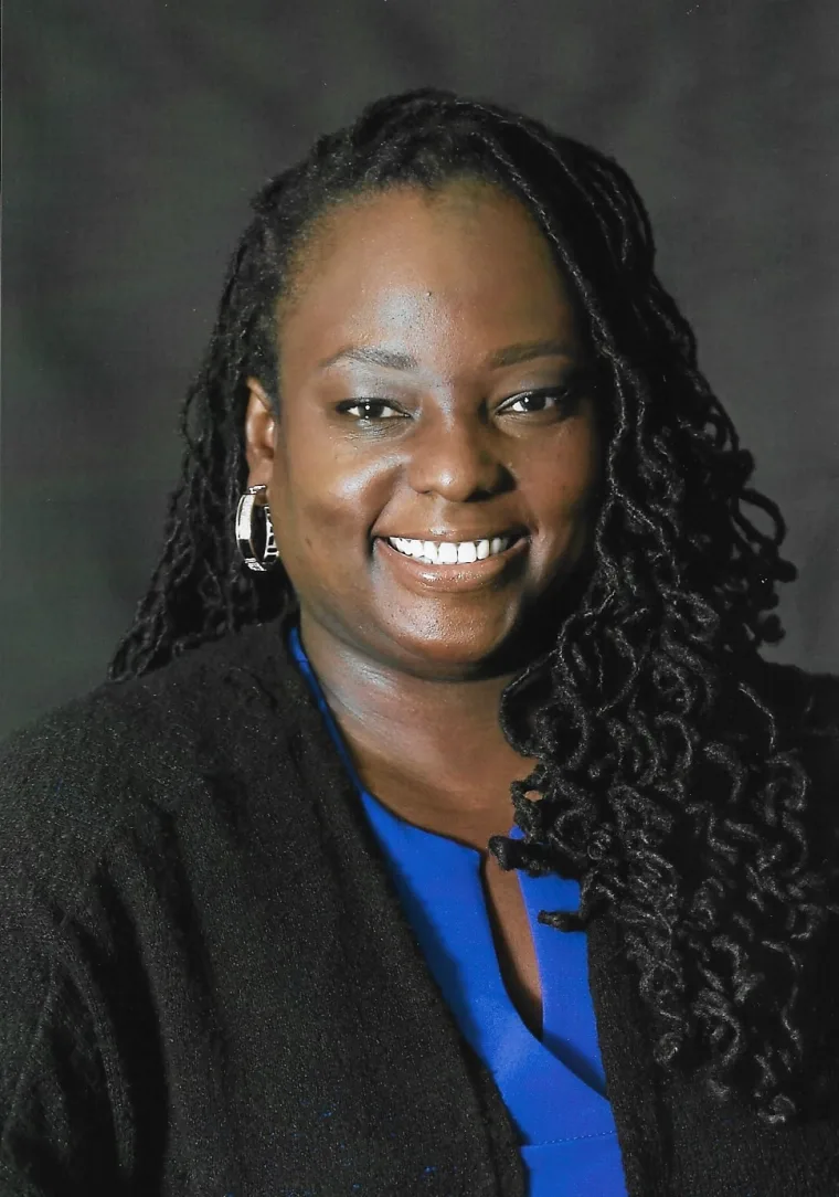 A person with long curly hair smiles, wearing a blue shirt and black jacket against a dark background.