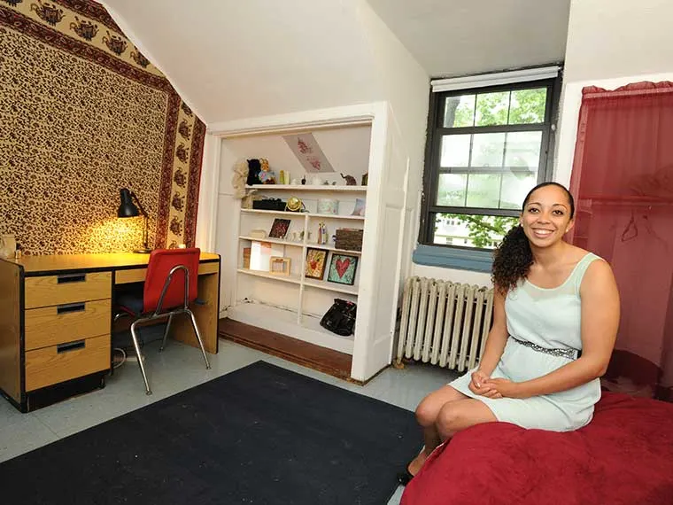 Person sitting on a bed in a cozy room with a tapestry, desk, shelves, and a window.