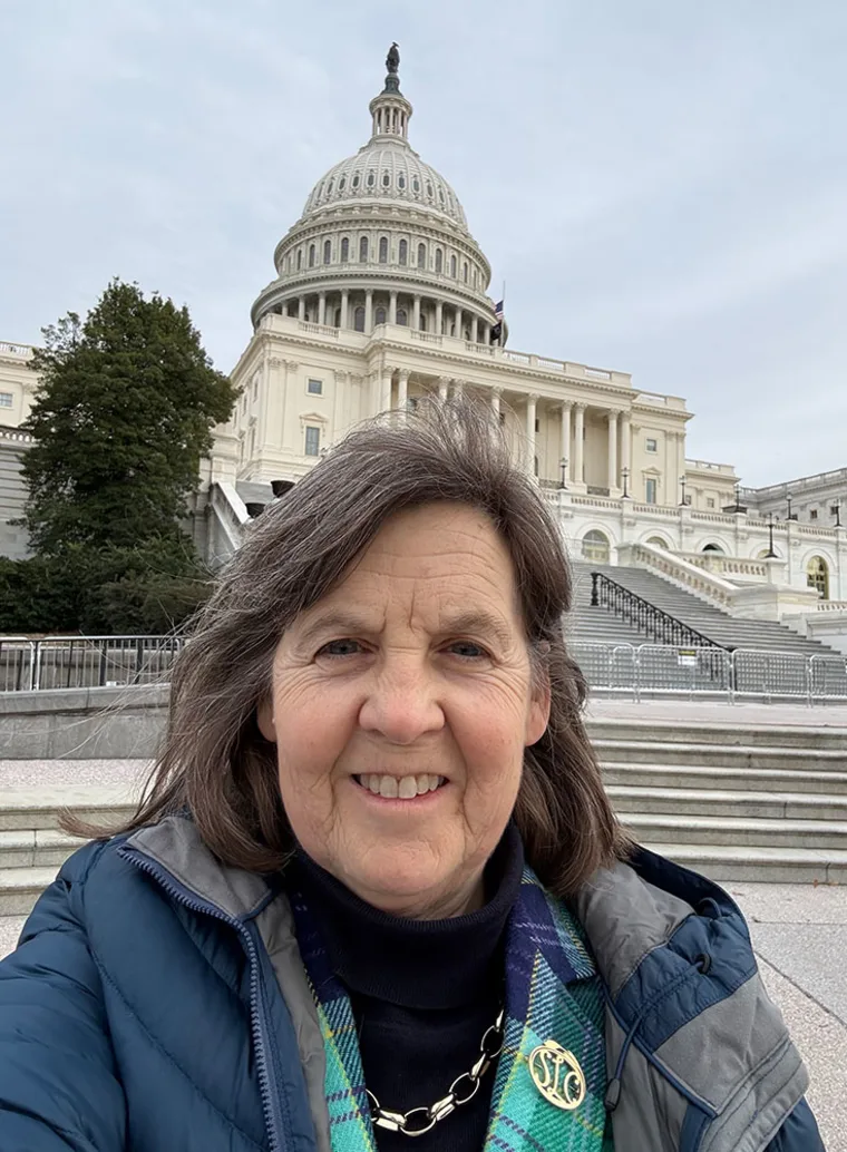 Person smiling in front of the U.S. Capitol building, wearing a blue jacket and scarf. Overcast sky visible in the background.