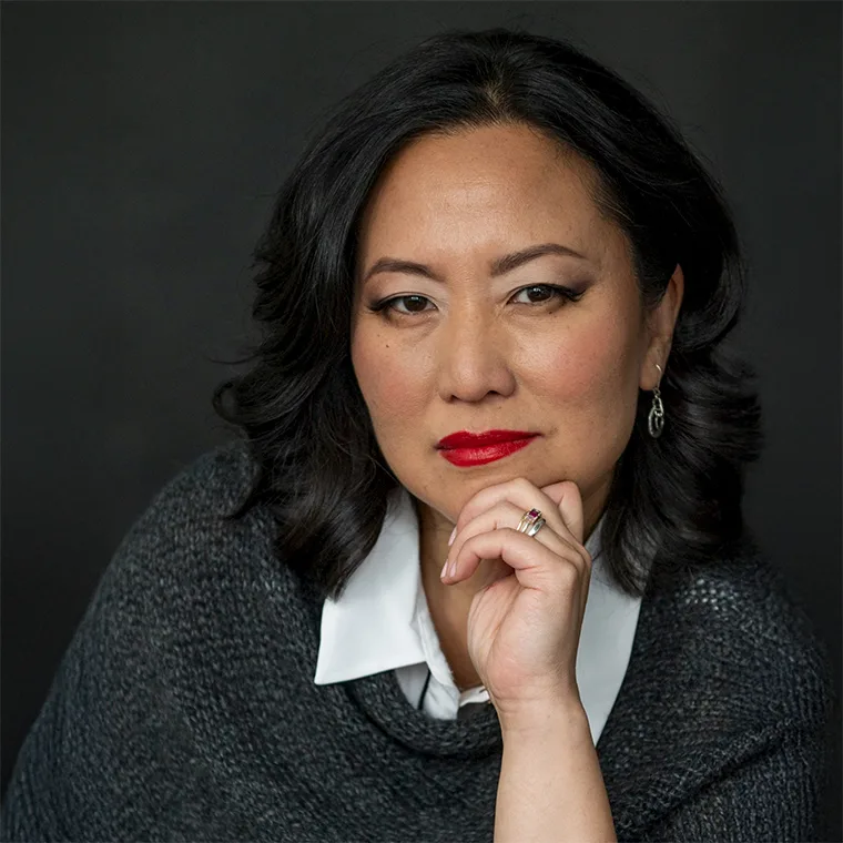 Person with dark hair, wearing a gray sweater and white collared shirt, resting chin on hand, with a thoughtful expression.