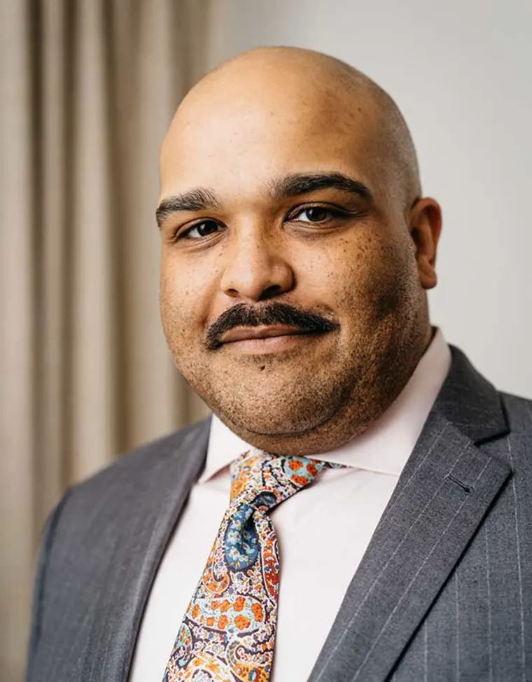 Person with a shaved head and mustache wearing a suit and a colorful tie, standing in front of a curtain.