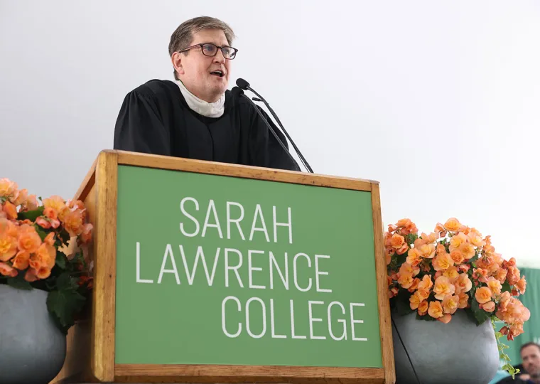 Person in academic robes speaks at a podium labeled "Sarah Lawrence College," with orange flowers on either side.