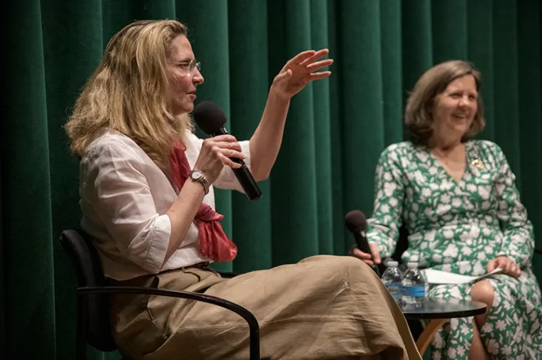 Two people sit onstage with microphones, engaged in a discussion. One gestures with a hand while the other sits, smiling. Green curtain backdrop.