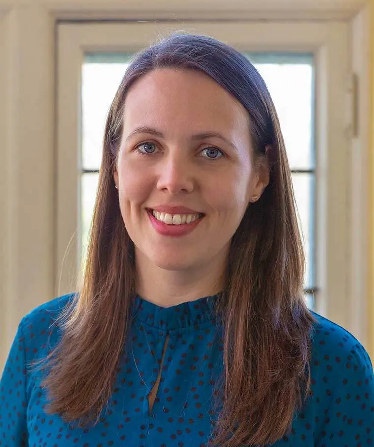 Person smiling, wearing a blue patterned top, standing indoors with a window in the background.