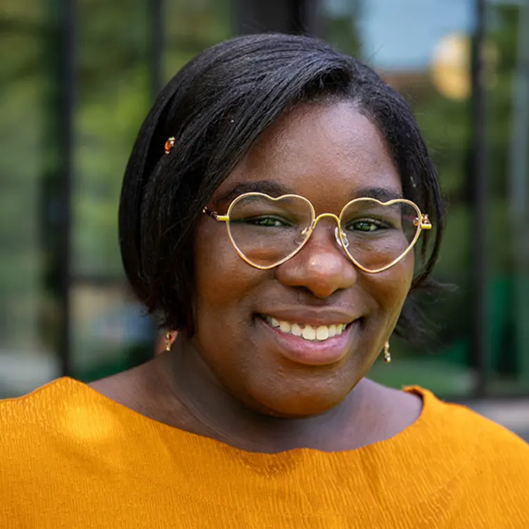 Person with short hair, wearing gold heart-shaped glasses and an orange top, smiling in front of a blurred background.