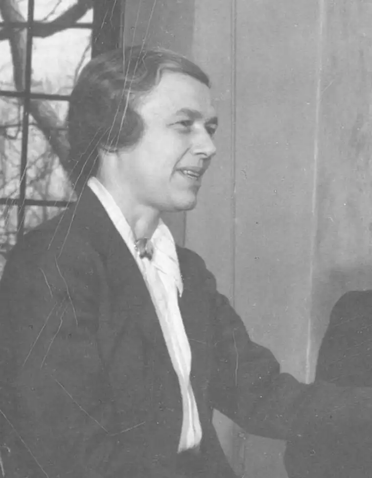 Person in a suit smiling, sitting near a window with tree branches visible outside. The photo is black and white and appears vintage.