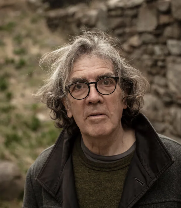 Person with gray hair and glasses, wearing a dark coat and green shirt, standing outdoors with a stone wall background.