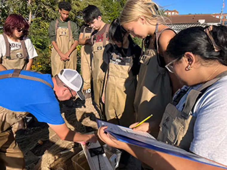 A group wearing aprons gathers around a wooden structure outdoors. One person writes on a clipboard while another points to the structure.