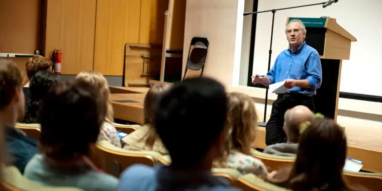 Person in blue shirt speaking to an attentive audience in an auditorium with a podium and microphone.