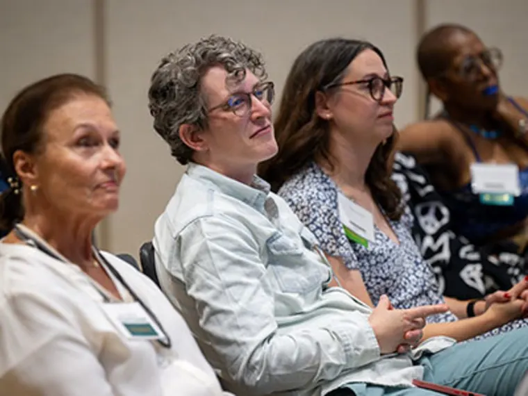 Four people seated in a conference setting, attentively listening to a presentation, with badges hanging from their necks.