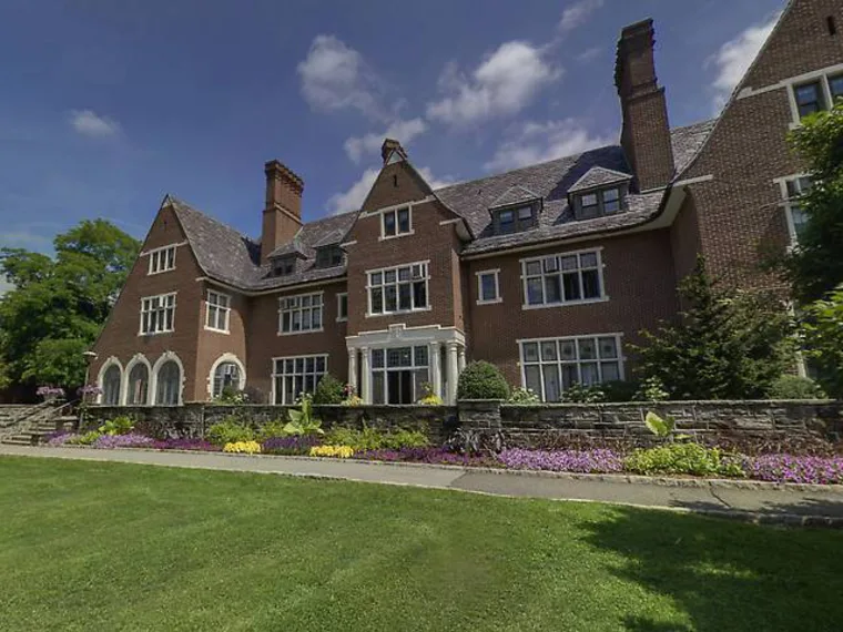 Large brick mansion with multiple chimneys, surrounded by manicured lawn and vibrant flowerbeds under a partly cloudy sky.