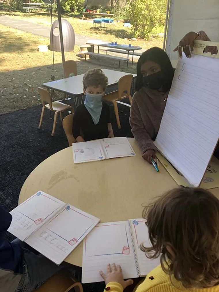 Children seated around a table with notebooks, one person showing a large pad, outdoors with playground equipment visible.