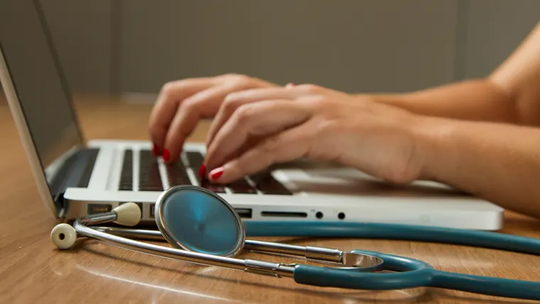 Hands typing on a laptop keyboard with a stethoscope on the wooden desk nearby.