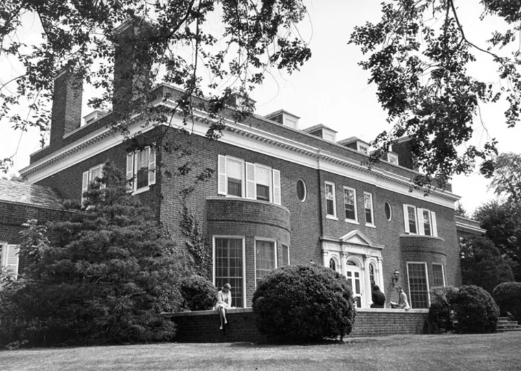 Historic brick building with large windows and chimneys, surrounded by trees and bushes. People are sitting and standing on the front steps.