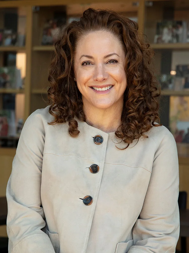 Person with curly hair smiling, wearing a light-colored coat with large buttons. Bookshelves are visible in the background.