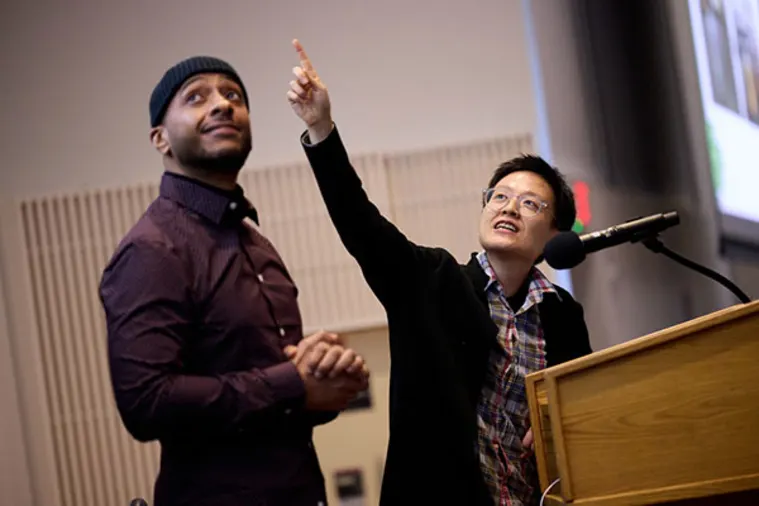 Two people stand near a podium. One points upwards while speaking into a microphone, and the other looks in the same direction with hands clasped.