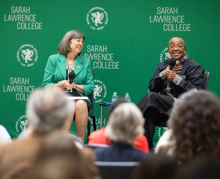 Two people seated with microphones on stage, smiling in discussion at Sarah Lawrence College, surrounded by an audience.