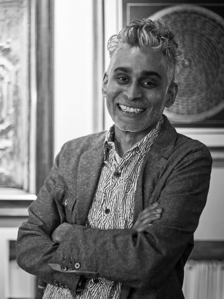 Person with short curly hair smiling, wearing a patterned shirt and blazer. Arms crossed. Art piece visible in the background. Black and white image.